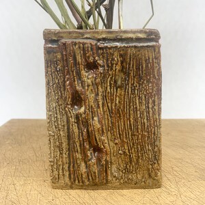 May include: A rectangular, textured ceramic vase with a brown, wood-like appearance. The vase contains dried flowers and stems. The vase is sitting on a light brown surface.
