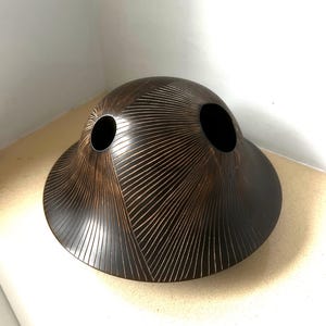 May include: A dark brown, dome-shaped vessel with a wide, flared base and two circular openings on the top. The surface features a repeating pattern of fine, light-coloured etched lines that radiate outwards in triangular sections across the exterior.