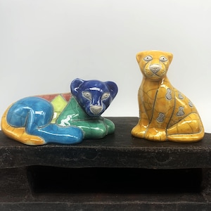 May include: Two ceramic animal figurines. One is a blue, green, and yellow reclining cat. The other is a yellow and gray sitting cat. Both have detailed patterns and are displayed on a dark wooden surface.