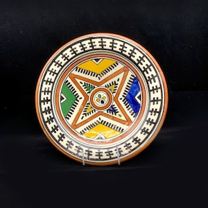 May include: A round ceramic plate featuring a colourful geometric design. The plate has a white background with a central star shape in orange, yellow, green, and blue. The border of the plate is a repeating pattern of black and white lines.