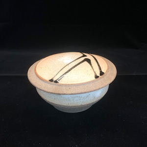 May include: A small, white ceramic bowl with a lid. The lid features a black abstract design. The bowl has a light brown rim and a white base.