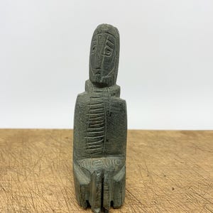 May include: A carved stone sculpture of a seated figure with a stylized face and beard. The figure is dark grey-green and has etched details on the chest and head. The sculpture is on a wooden surface.
