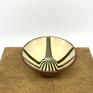 May include: A ceramic bowl with a cream interior and a dark brown rim. The bowl has a black, linear design that fans out from the centre. The bowl is set on a wooden surface.