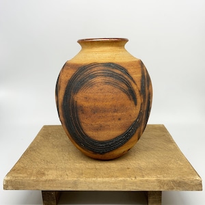May include: A handmade ceramic vase with a rounded body and a narrow neck. The vase has a warm, earthy colour palette with a brown base and a lighter tan top. Bold, black, curved lines encircle the vase, adding a unique design element.