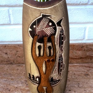 May include: A tall, cylindrical ceramic vase with a textured, metallic gold finish and a dark brown rim. The front features a relief design of a large stylised fish, two smaller vertical fish, and a small boat, all rendered in shades of brown.