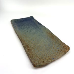 May include: Rectangular ceramic serving tray with a textured surface. The tray has a gradient of blue and green hues, with a darker blue at the top and a speckled, earthy green at the bottom. The edges have a rustic, brown finish.