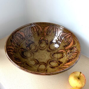 May include: A handmade ceramic bowl with a wide, shallow design. The bowl has a speckled, earthy green base with a repeating pattern of brown and orange floral-like designs. Suitable for serving or display. Measures approximately 30cm in diameter.