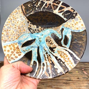May include: A ceramic plate with an abstract design in brown, black, white and blue. The plate has a raised rim and a slightly uneven surface.