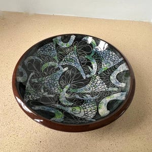 May include: A ceramic bowl with a dark brown rim and a black interior. The bowl has a detailed pattern of white, blue, and green abstract shapes. The bowl is likely used for serving or display.
