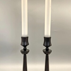 Pode incluir: Um par de casti&ccedil;ais de madeira preta com velas brancas. Os casti&ccedil;ais t&ecirc;m um design simples e elegante com uma base redonda e um caule alto e fino.