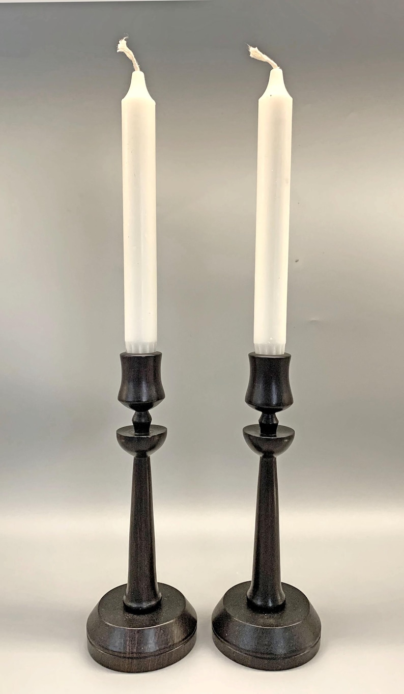 Pode incluir: Um par de casti&ccedil;ais de madeira preta com velas brancas. Os casti&ccedil;ais t&ecirc;m um design simples e elegante com uma base redonda e um caule alto e fino.