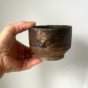 May include: Handmade ceramic cup with a textured, earthy appearance. The cup features a dark brown exterior with lighter brown and gold accents near the rim. The cup has a slightly flared shape and a rough, tactile surface.