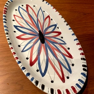 May include: White oval ceramic serving dish with a blue and red floral design. The design features a stylised flower with black dots and thin lines.