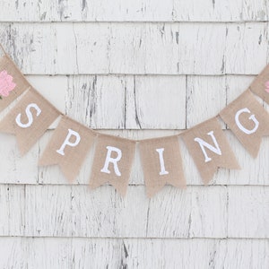 May include: A burlap banner with the word "SPRING" spelled out in white letters. The banner is decorated with two pink flowers.