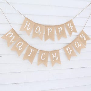 May include: A banner with the words "Happy Match Day" written in white script on individual tan pennants. The banner is strung on twine and hangs against a white wooden background.