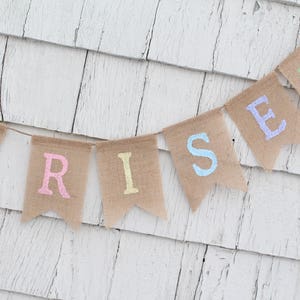 Może przedstawiać: Baner z pł&oacute;tna jutowego z napisem "RISEN" napisanym pastelowymi kolorami. Każda litera znajduje się na osobnej fladze. Baner wisi na białej drewnianej ścianie.