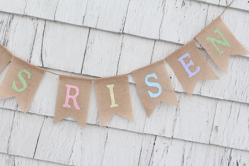 Może przedstawiać: Baner z pł&oacute;tna jutowego z napisem "RISEN" napisanym pastelowymi kolorami. Każda litera znajduje się na osobnej fladze. Baner wisi na białej drewnianej ścianie.