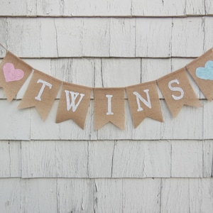 May include: A burlap banner with the word "TWINS" in white letters. The banner is decorated with a pink heart on the left and a blue heart on the right.