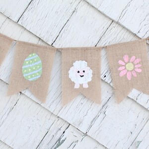 K&ouml;nnte beinhalten: Ein Burlap-Banner mit bemalten Ostern-Motiven. Das Banner zeigt einen Hasen, ein Ei, ein Lamm, eine Blume und ein K&uuml;ken.