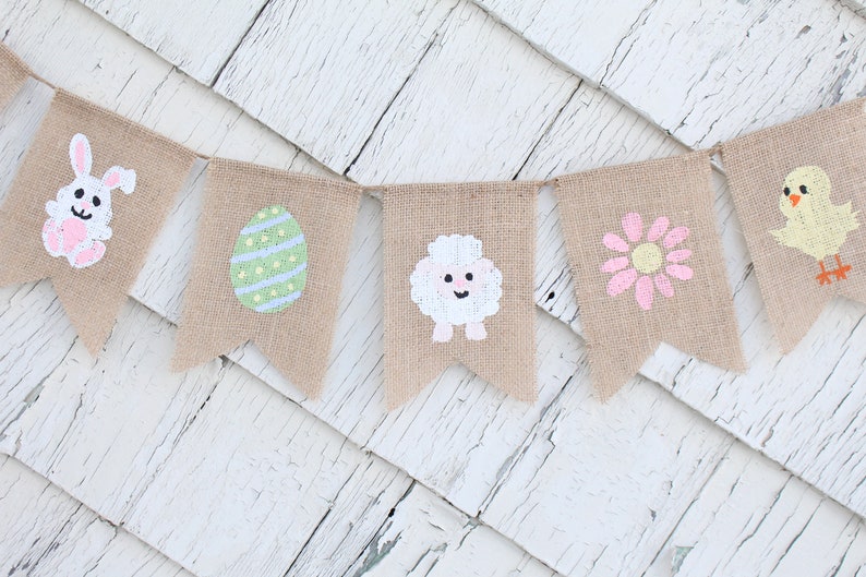 K&ouml;nnte beinhalten: Ein Burlap-Banner mit bemalten Ostern-Motiven. Das Banner zeigt einen Hasen, ein Ei, ein Lamm, eine Blume und ein K&uuml;ken.