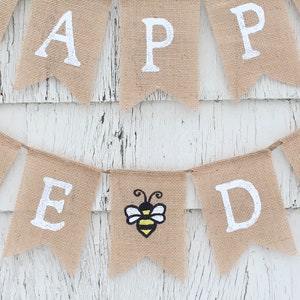 May include: A burlap banner with the words "Happy Bee Day" in white paint. A small bee with yellow and black stripes is in the center of the banner.