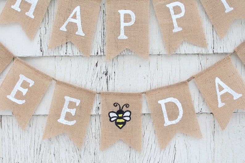 May include: A burlap banner with the words "Happy Bee Day" in white paint. A small bee with yellow and black stripes is in the center of the banner.
