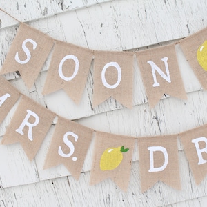 May include: A burlap banner with the words "Soon to Mrs." and a lemon graphic. The banner is strung on a white wooden background.