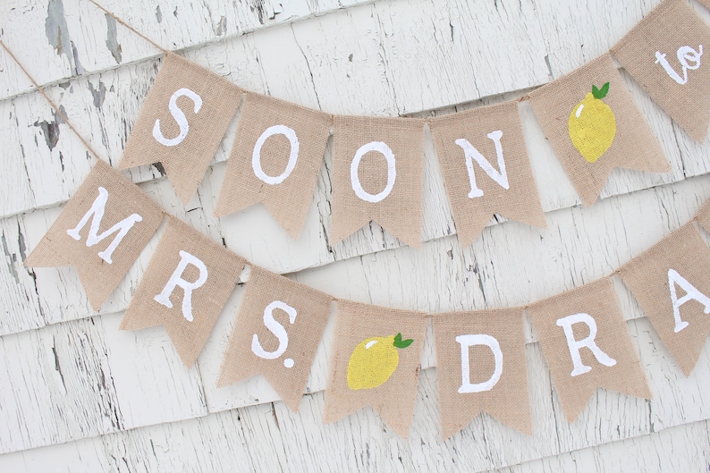 May include: A burlap banner with the words "Soon to Mrs." and a lemon graphic. The banner is strung on a white wooden background.