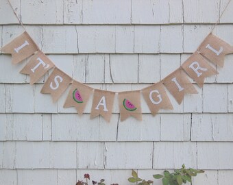 May include: A burlap banner with the words "It's a Girl" in white paint. The banner is decorated with watermelon slices.