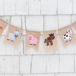 May include: A burlap banner with farm animal illustrations. The banner features a cow, rooster, goat, pig, horse, and another cow. The animals are painted in various colors, including white, black, brown, red, orange, and pink.