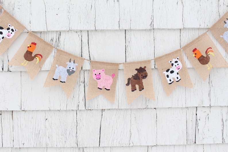May include: A burlap banner with farm animal illustrations. The banner features a cow, rooster, goat, pig, horse, and another cow. The animals are painted in various colors, including white, black, brown, red, orange, and pink.