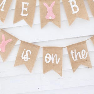 May include: A burlap banner with pink bunny cutouts and white lettering that reads "Baby is on the way".