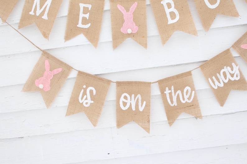 May include: A burlap banner with pink bunny cutouts and white lettering that reads "Baby is on the way".