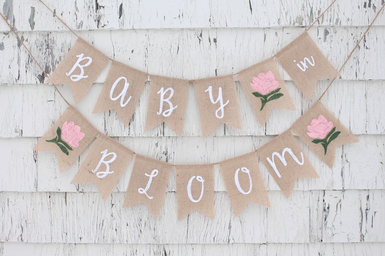 K&ouml;nnte beinhalten: Ein Banner aus Jute mit den Worten "Baby in Bloom" in wei&szlig;er Kursivschrift. Das Banner ist mit rosa Blumen und gr&uuml;nen Bl&auml;ttern verziert.