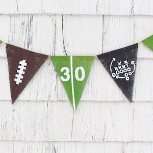 Könnte beinhalten: Ein grünes und braunes Burlap-Banner mit Fußball-Dekorationen. Das Banner hat die Zahlen 20, 30 und 40, einen Fußball und ein Football-Spieldiagramm.