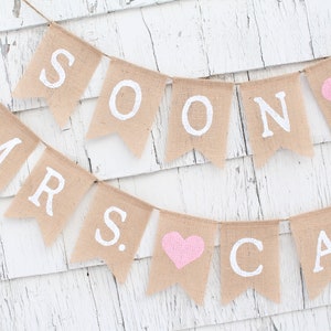 May include: A burlap banner with the words "Soon to be Mrs." in white paint. The banner has pink hearts between the words.