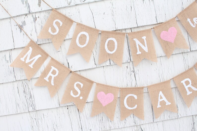 May include: A burlap banner with the words "Soon to be Mrs." in white paint. The banner has pink hearts between the words.
