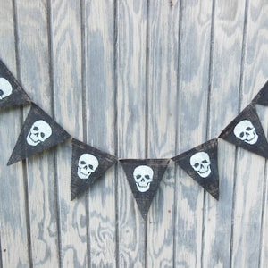May include: A black burlap banner with white skull designs. The banner is made of triangular flags strung together with twine. The skulls are hand-painted and add a spooky touch to the Halloween decor.