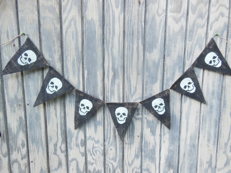 May include: A black burlap banner with white skull designs. The banner is made of triangular flags strung together with twine. The skulls are hand-painted and add a spooky touch to the Halloween decor.