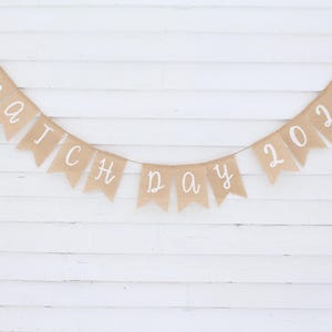 May include: A banner made of tan burlap flags strung together with white text that reads "match day 2026". The banner is hanging against a white wooden background.