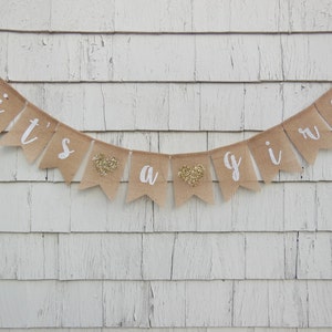 May include: A burlap banner with the words "it's a girl" in white paint. The banner has gold glitter hearts between the words.