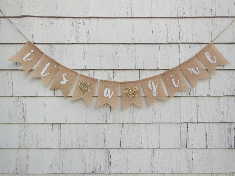 May include: A burlap banner with the words "it's a girl" in white paint. The banner has gold glitter hearts between the words.