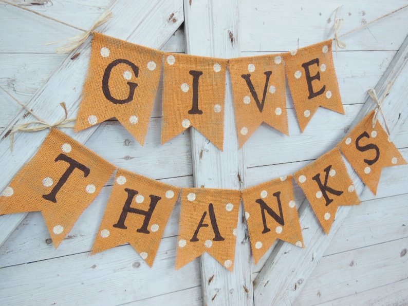 May include: A rustic orange burlap banner with the words "GIVE THANKS" in dark brown letters. Each pennant has white polka dots and is strung together with twine, perfect for Thanksgiving or fall decor.