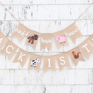 Pu&ograve; includere: Un banner di juta con il nome "JACK IS TWO" in lettere bianche. Include illustrazioni di animali e suoni come "oink" e "muu". Lo sfondo &egrave; in legno bianco invecchiato.