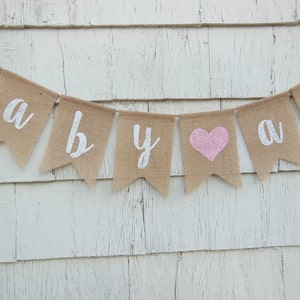 May include: A burlap banner with the words "baby" in white letters and a pink heart in the center. The banner is strung on a white wooden wall.