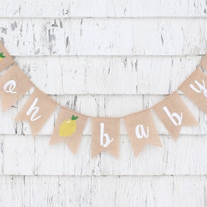 Peut inclure: Une banni&egrave;re en toile de jute avec les mots "oh baby" peints en blanc. Chaque lettre est sur un drapeau s&eacute;par&eacute; avec une illustration de citron jaune. La banni&egrave;re est accroch&eacute;e &agrave; un mur en bois blanc.