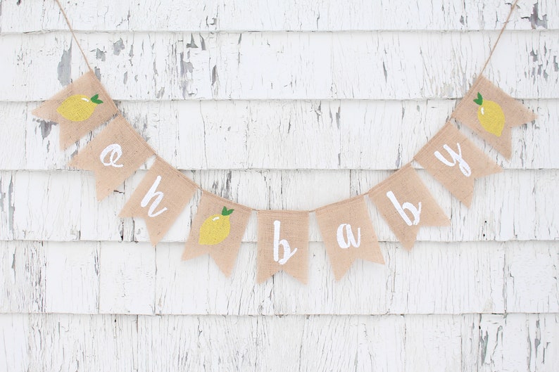 Peut inclure: Une banni&egrave;re en toile de jute avec les mots "oh baby" peints en blanc. Chaque lettre est sur un drapeau s&eacute;par&eacute; avec une illustration de citron jaune. La banni&egrave;re est accroch&eacute;e &agrave; un mur en bois blanc.