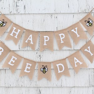 May include: A burlap banner with the words "Happy Bee Day" in white letters. The banner is decorated with three yellow and black bees.
