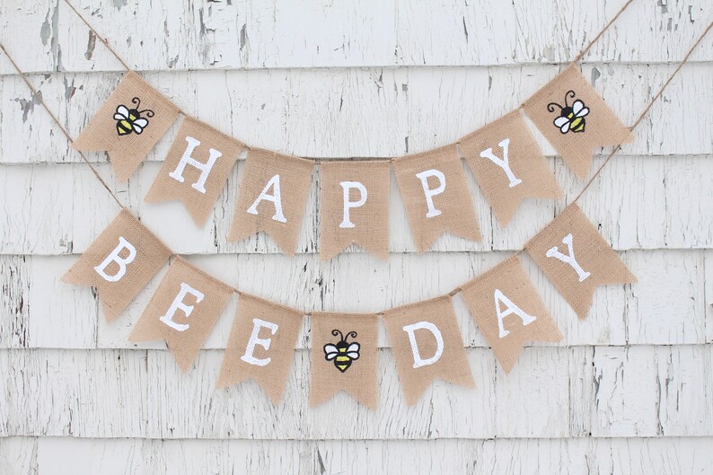 May include: A burlap banner with the words "Happy Bee Day" in white letters. The banner is decorated with three yellow and black bees.