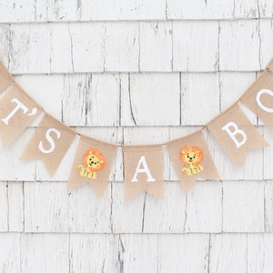May include: A burlap banner with the words "It's a Boy" in white letters. The banner is decorated with small lion illustrations.